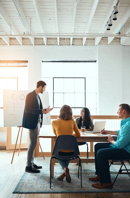 Mężczyzna w garniturze przedstawia prezentację trzem współpracownikom siedzącym przy stole w jasnym biurze. Wyglądają na zaangażowanych, a w tle widać duże okno i tablicę flipchart