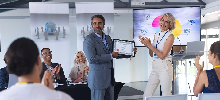 Podczas konferencji mężczyzna otrzymuje certyfikat od uśmiechniętej kobiety na scenie. Publiczność oklaskuje. Za nimi widoczny jest kolorowy wykres kołowy