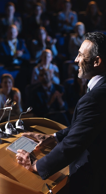 Mężczyzna w garniturze przemawia pewnie z podium wyposażonego w mikrofony i tablet cyfrowy, zwracając się do zafascynowanej, oklaskującej publiczności w słabo oświetlonym pomieszczeniu