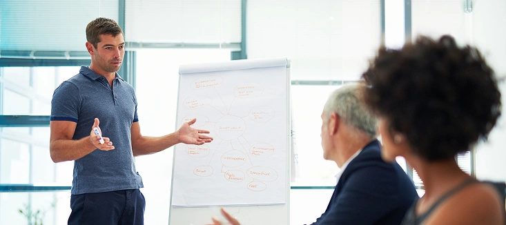 Mężczyzna w niebieskiej koszuli prezentuje diagramy na tablicy flipchart, angażując dwóch kolegów siedzących przy stole. Otoczenie jest jasne i profesjonalne