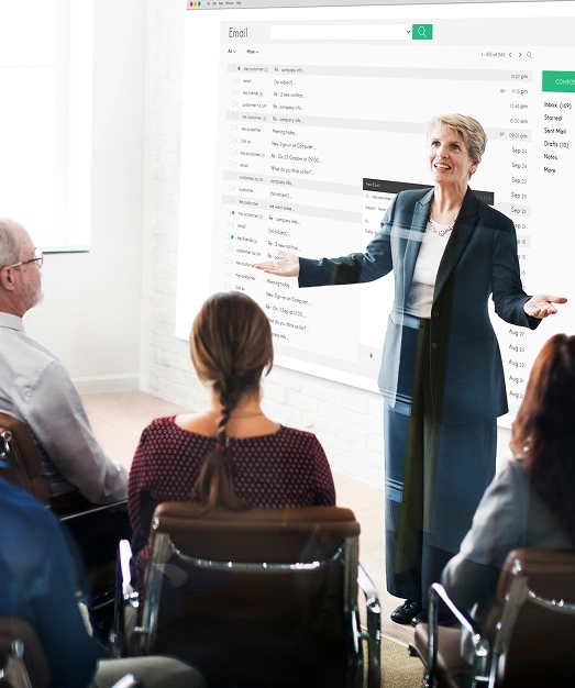 Kobieta entuzjastycznie prezentuje się grupie osób siedzących w biurze. Duży wyświetlany interfejs poczty elektronicznej służy jako tło, wskazując na spotkanie poświęcone technologii