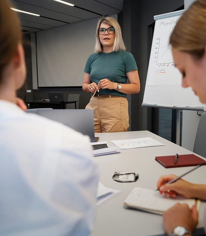Kobieta w zielonej koszuli pewnie prowadzi spotkanie biznesowe, wskazując gestem na tablicę z wykresami. Dwie osoby uważnie robią notatki