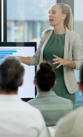 Profesjonalna kobieta w zielonej sukience i szarym blezerze prezentuje dane na ekranie przed uważną publicznością. Akcja rozgrywa się w nowoczesnym biurze z przeszklonymi ścianami