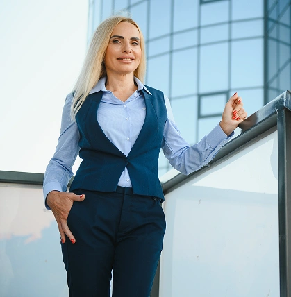 Pewna siebie kobieta w stroju biznesowym stoi na zewnątrz i uśmiecha się. Opiera się o szklaną balustradę, a za nią widać nowoczesny szklany budynek, co podkreśla jej profesjonalizm