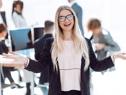 Uśmiechnięta kobieta o długich blond włosach i w okularach, ubrana w czarną marynarkę, stoi z otwartymi ramionami w jasnym biurze. W tle pracują koledzy